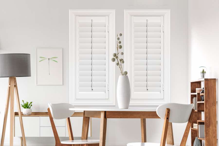 White walls and white Polywood shutters can give a room a fresh, minimalist aesthetic. White Polywood plantation shutters on two skinny windows in a white dining room