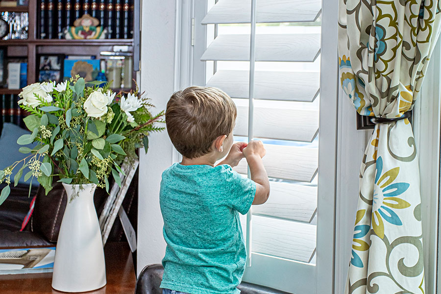 Cordless window shutters are a great safety feature for small children and pets. A little boy peaking through white polywood shutters